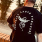 Man with tattoos sat on brick wall wearing black tshirt with a white print of text  saying Nothing Last Forever and a skeleton hand holding a dead rose