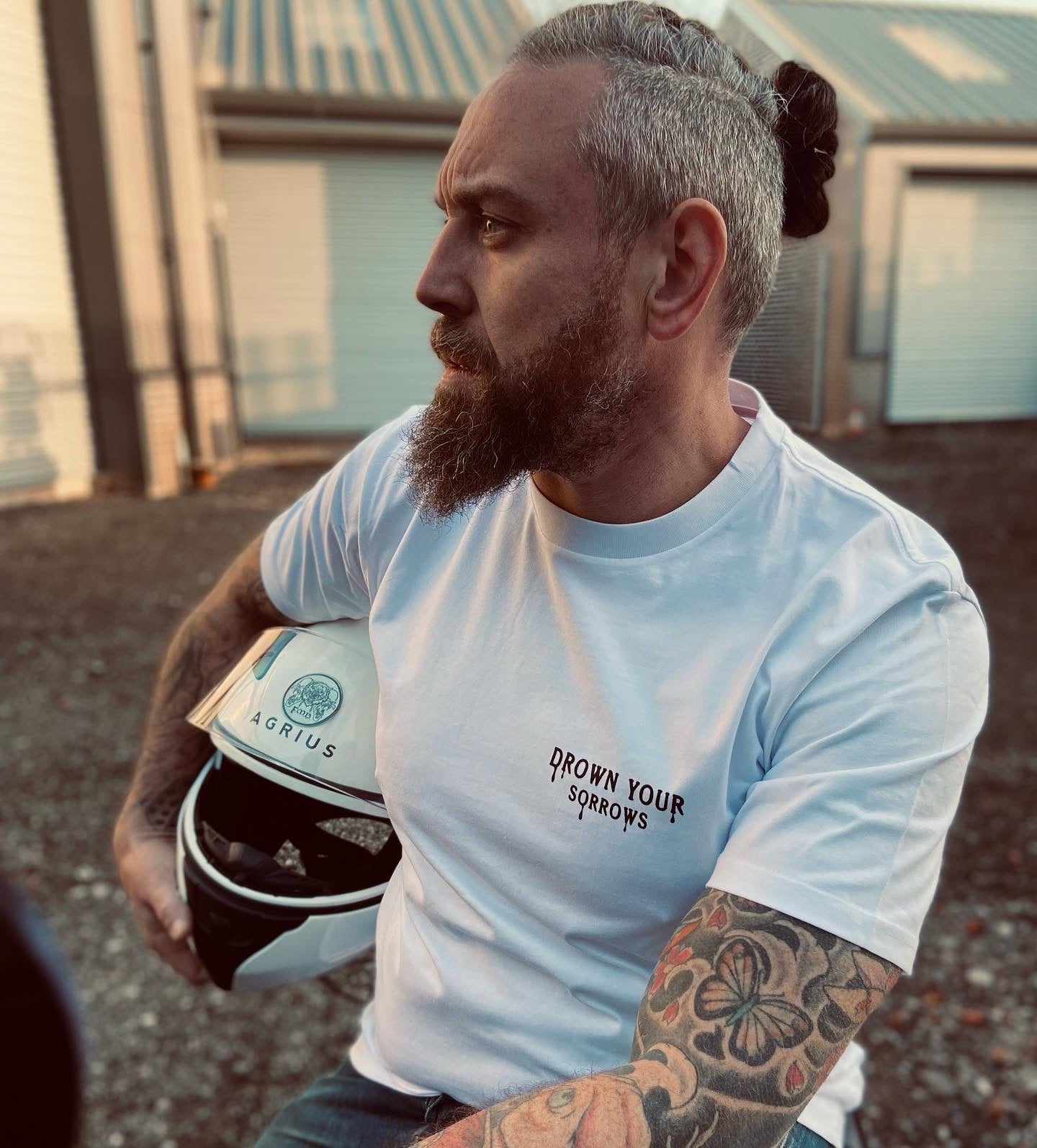 man with beard and tattoos holding motorbike helmet, wearing white tshirt with black print on left chest, Drown Your Sorrows