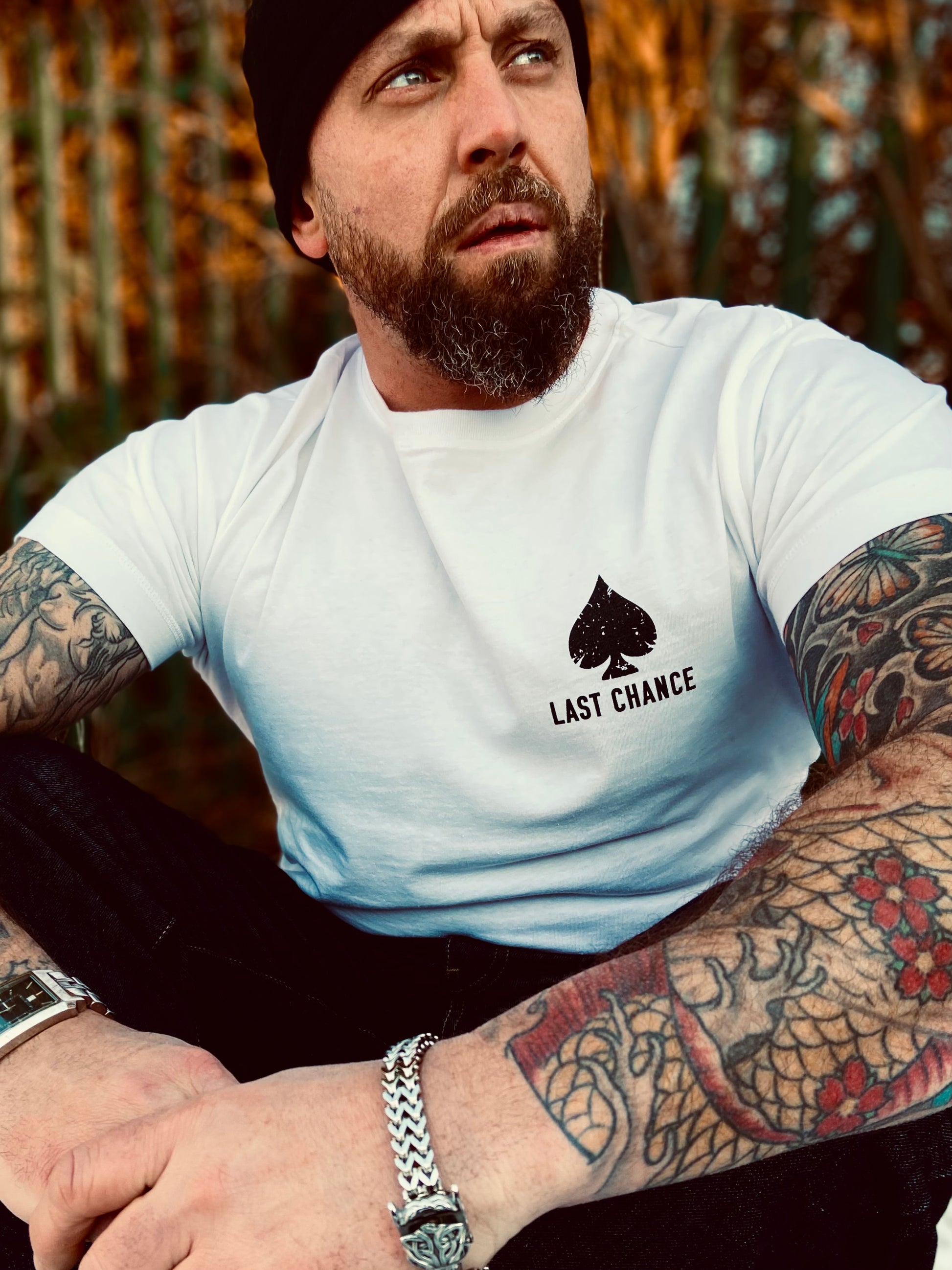 man with black hat, beard and tattoos sitting down, white tshirt with black print of playing card space, Last Chance text